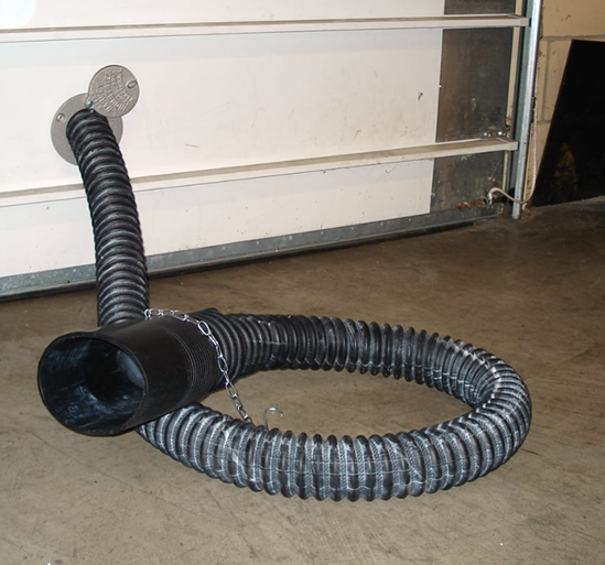 Black exhaust hose venting from a garage door, looped on the concrete floor. A metal plate surrounds the hose opening.