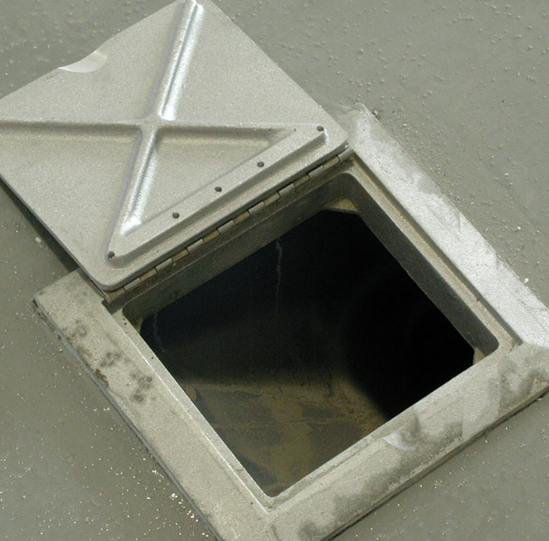 Open metal access hatch on a gray floor. The square lid is partially open, revealing a dark interior.