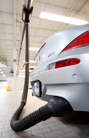 Silver car undergoing an emissions test. A black hose is attached to the exhaust pipe, collecting exhaust fumes in a garage.