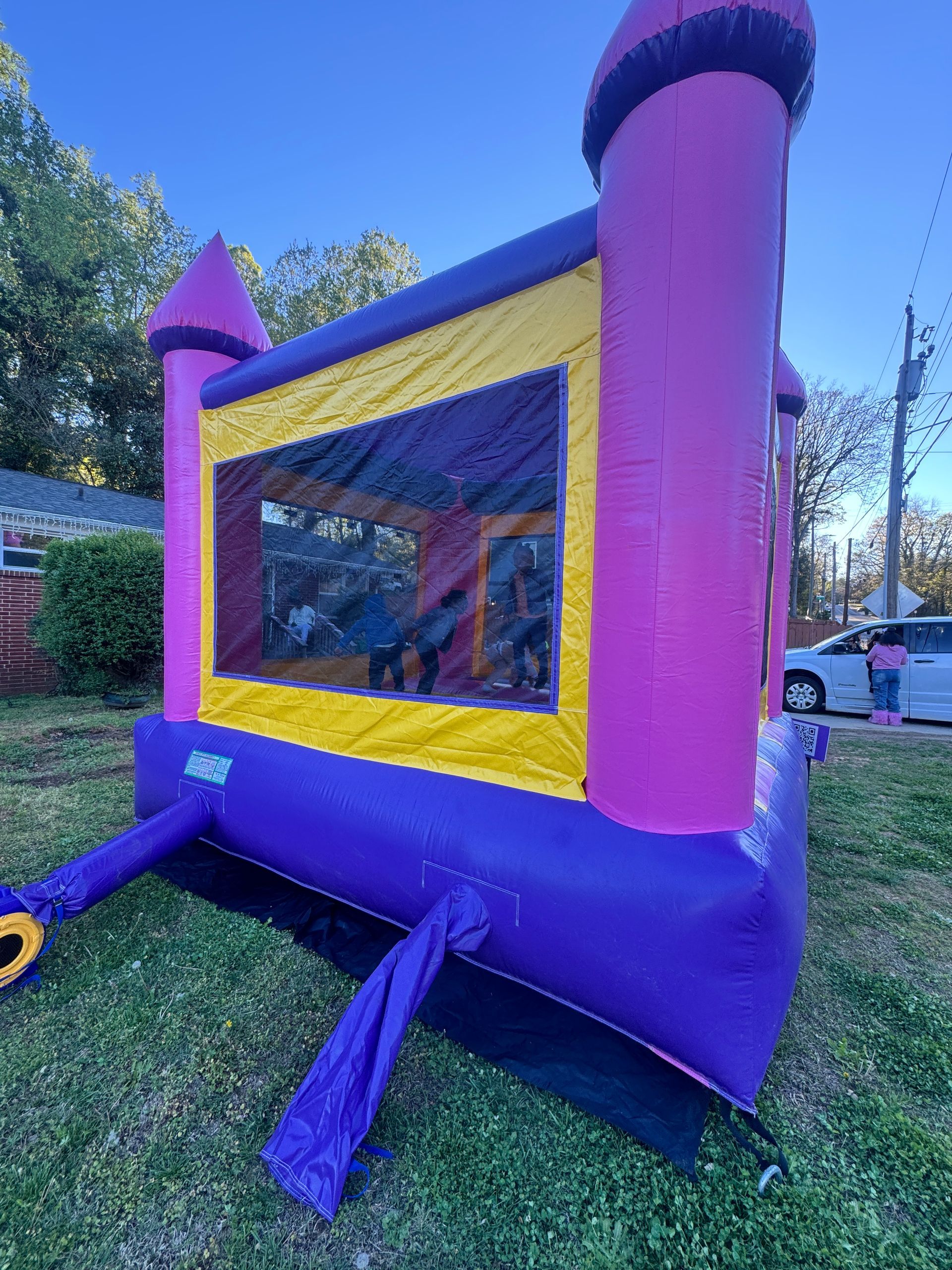 A bright pink and purple bouncy castle sits on a green lawn with a white car parked in the background.