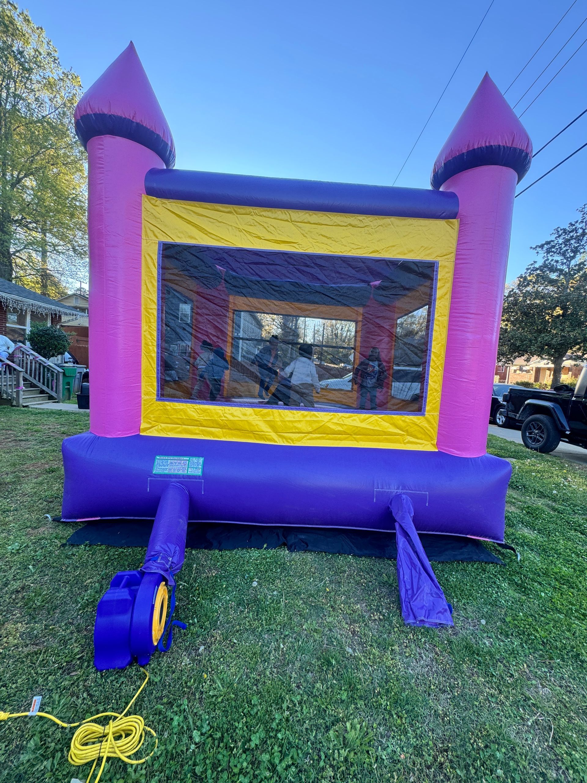 A pink and purple inflatable bounce house stands on a grassy lawn with a black blower unit attached in the front.