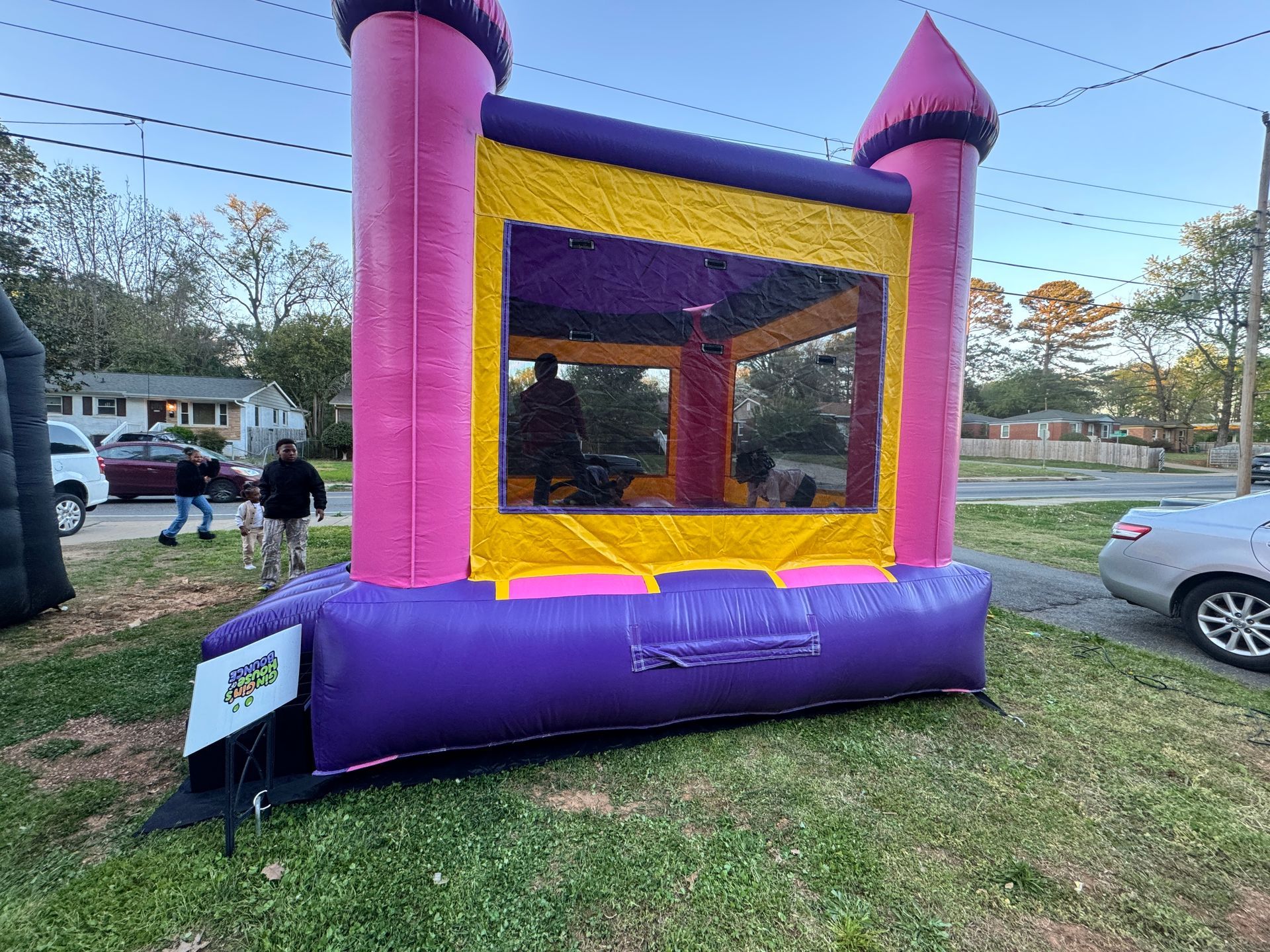 A pink and purple inflatable bounce house stands on a grassy lawn near a road with parked cars and people nearby.