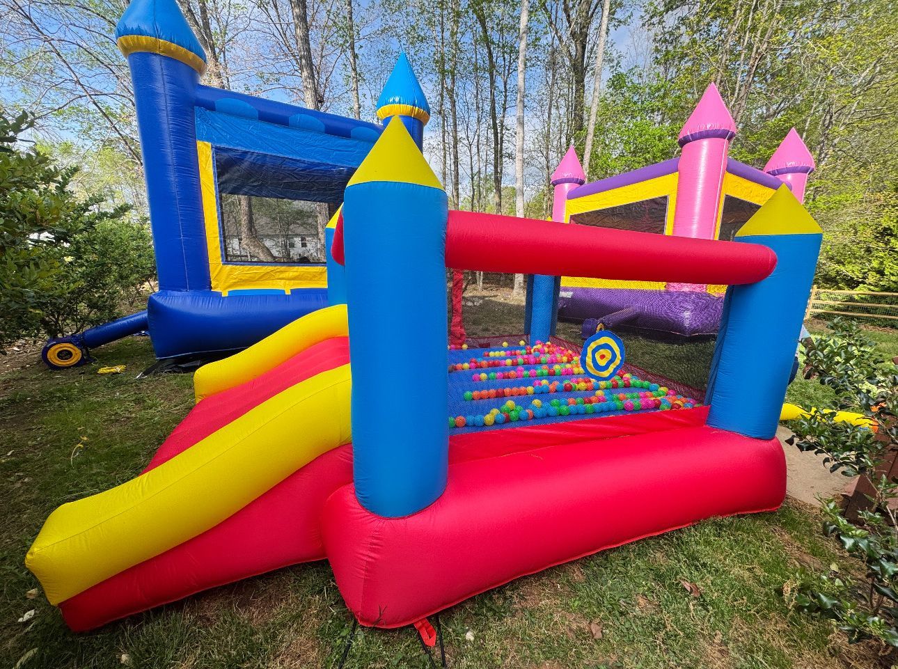 A colorful inflatable bounce house with a slide and a ball pit, situated on a grassy area outdoors.