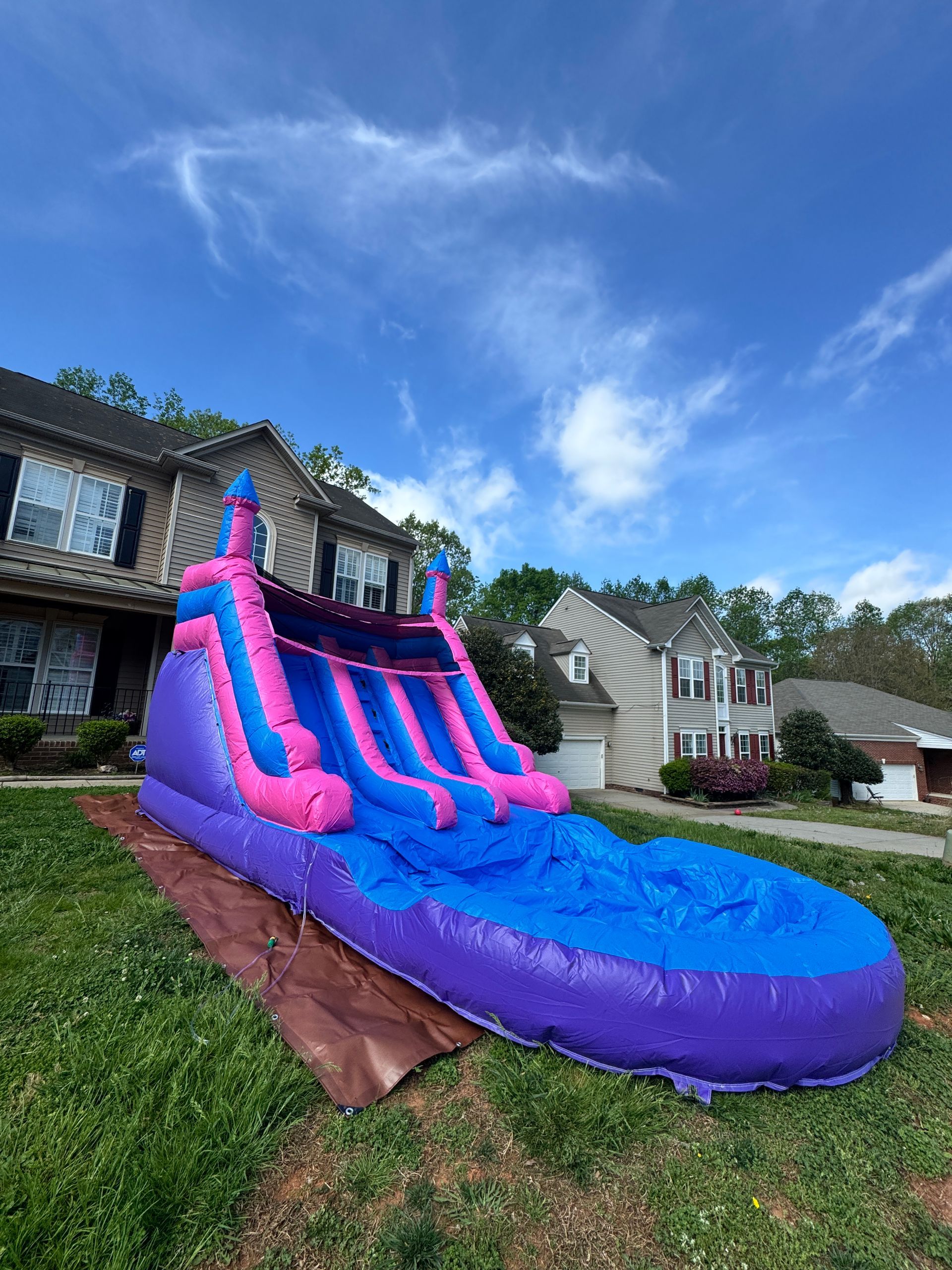 A large purple, pink, and blue inflatable water slide sits on a brown tarp in a suburban front yard.