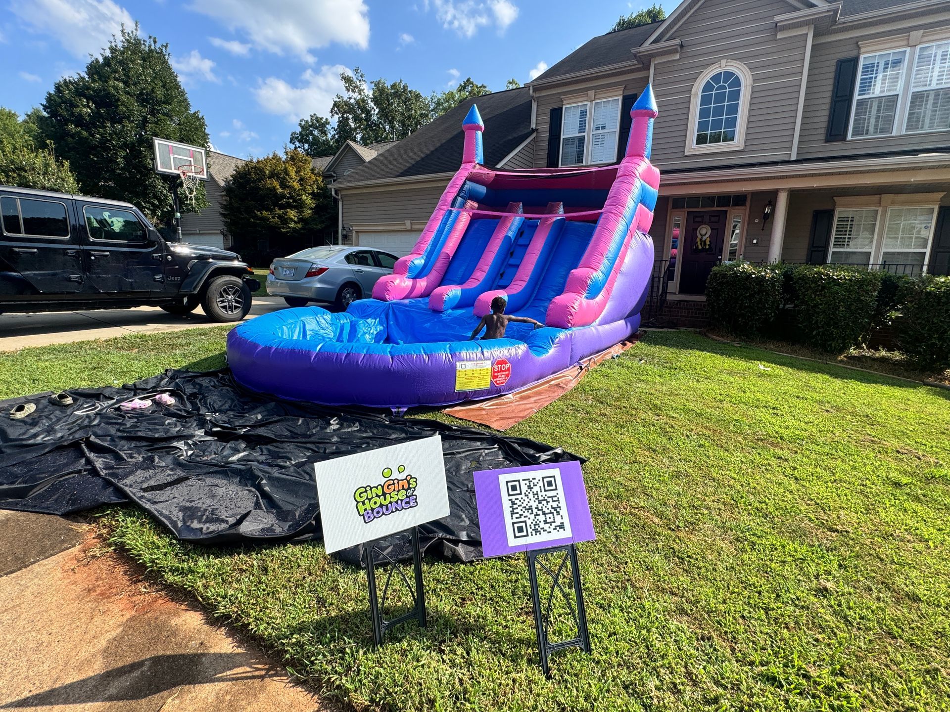 A pink and purple inflatable water slide sits on a residential lawn, with a small sign featuring a QR code in the foreground.