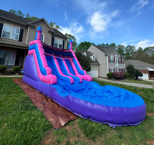 A large, dual-lane, purple and blue inflatable water slide set up on a grassy lawn in front of suburban houses.