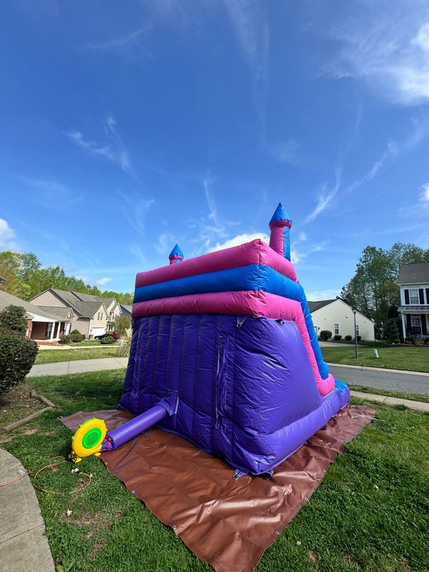 A purple, pink, and blue inflatable bounce house slide sits on a brown tarp in a grassy residential yard.