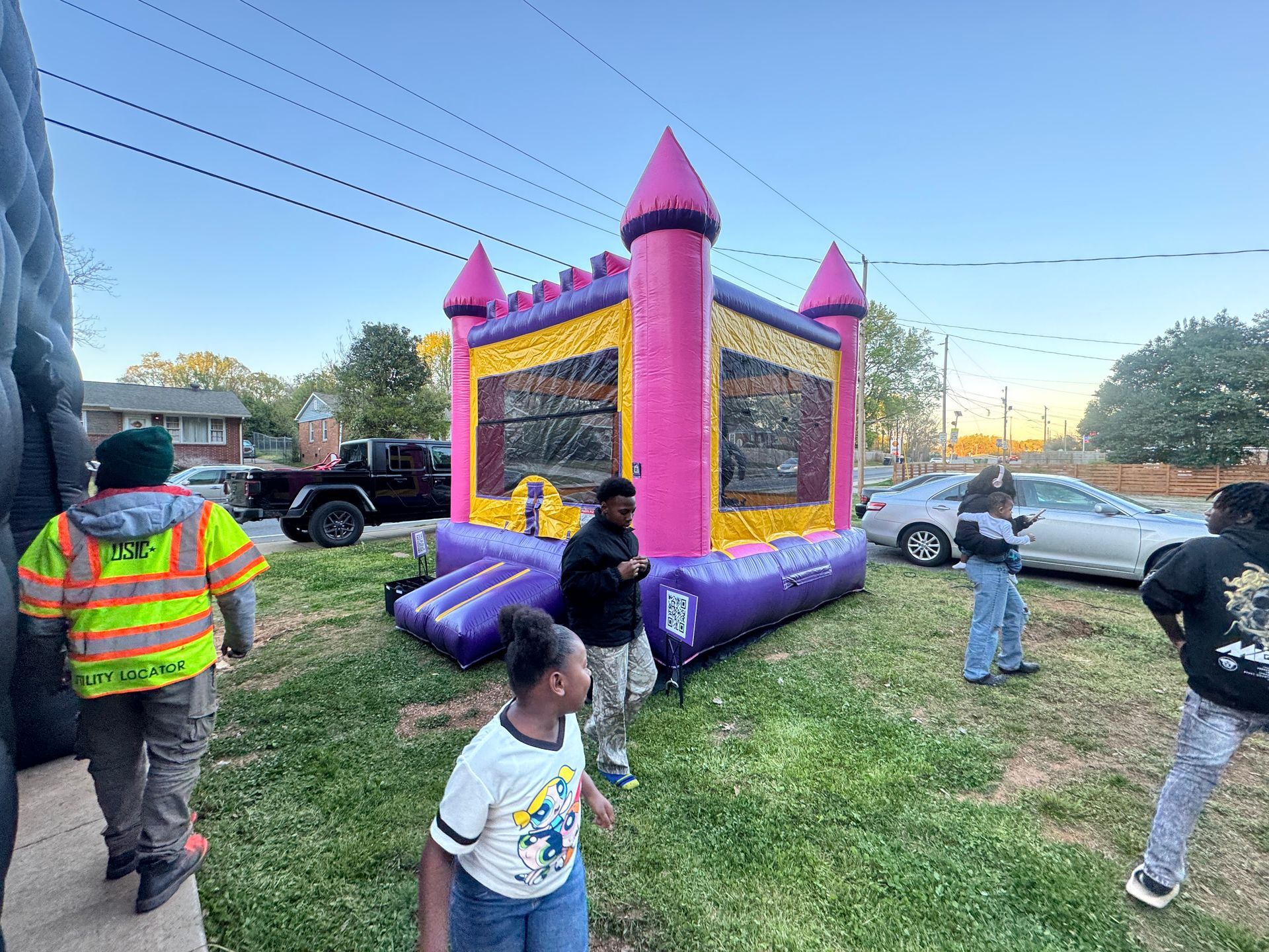 A vibrant pink and yellow bounce house stands in a grassy yard, surrounded by several people in casual clothing.