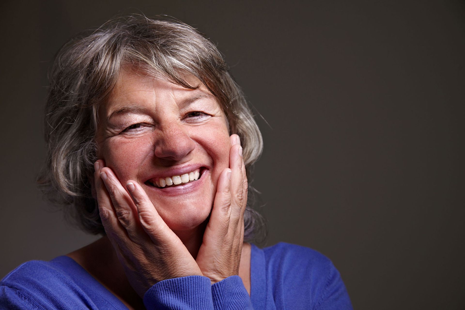 An older woman is smiling with her hands on her face.