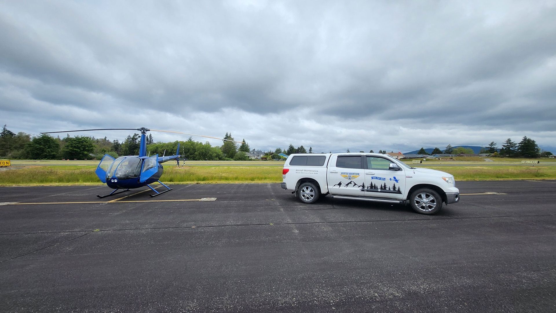 A blue helicopter on its side on a runway next to a white truck with logos under cloudy skies.