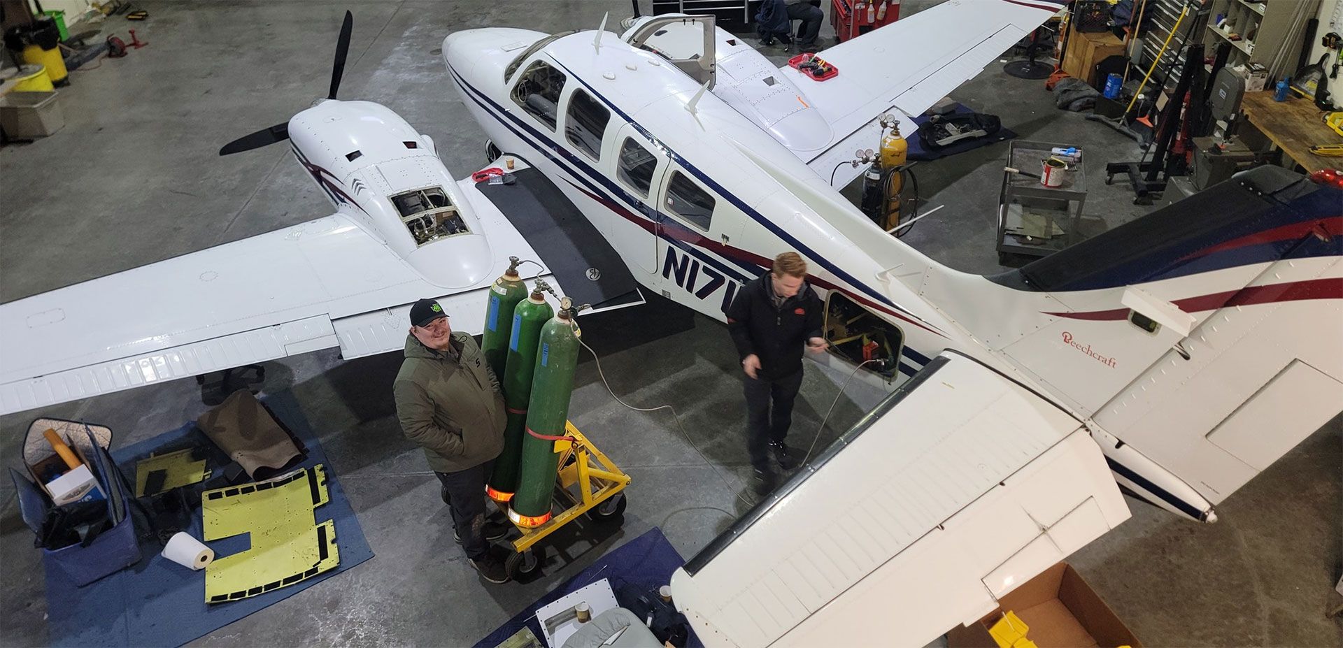 Two people near a plane in a hangar; one near oxygen tanks, the other at an opened panel.