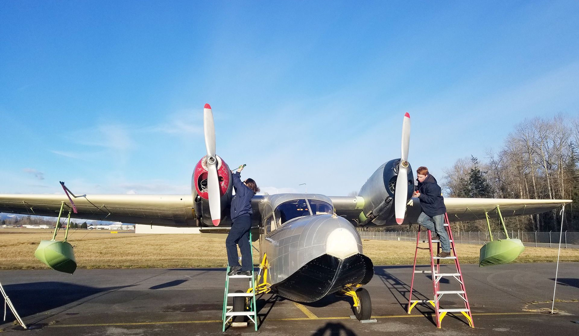 Two people work on a seaplane on a tarmac under a blue sky.