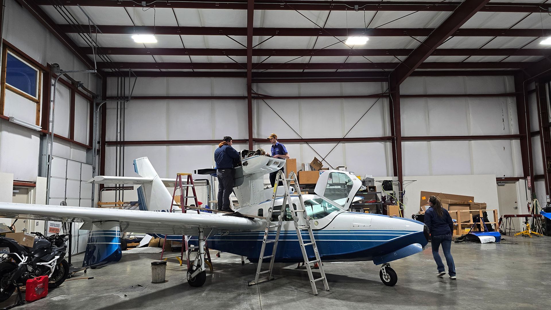 Workers assembling a seaplane inside a hangar with tools. White, blue seaplane. Men, woman.