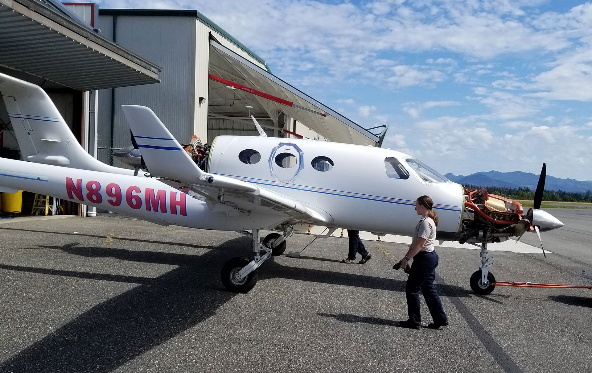 White airplane with pink lettering, mechanics working, parked on tarmac near an open hangar.