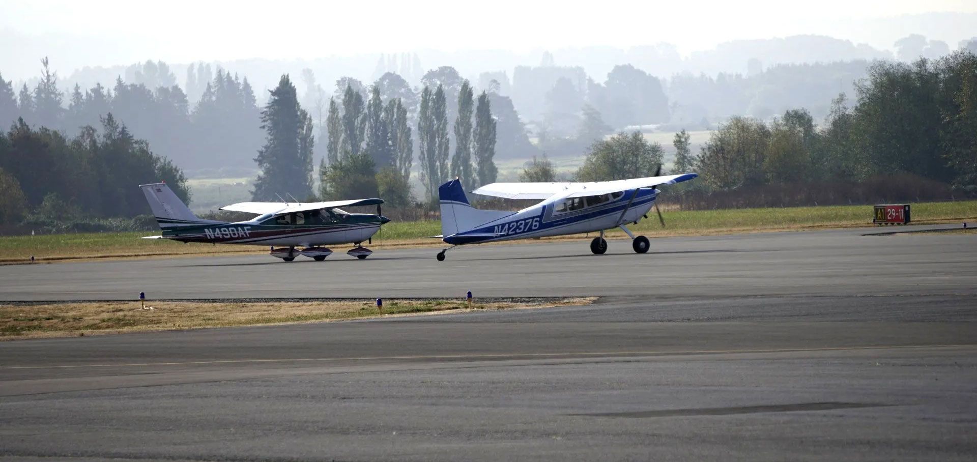 Two small airplanes on a gray tarmac; trees and a hazy sky in the background.