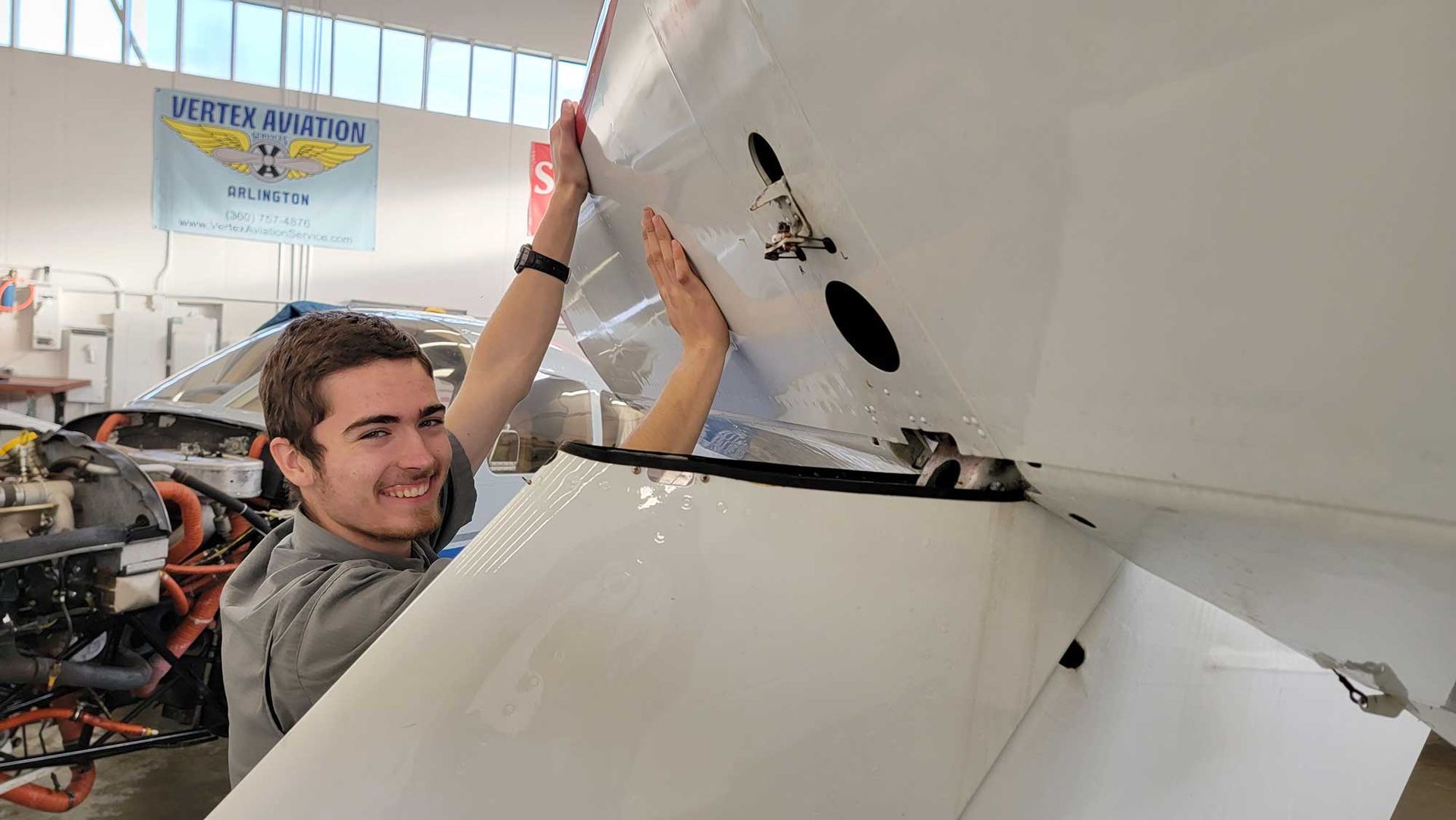 Man smiling, working on white aircraft wing inside a workshop.