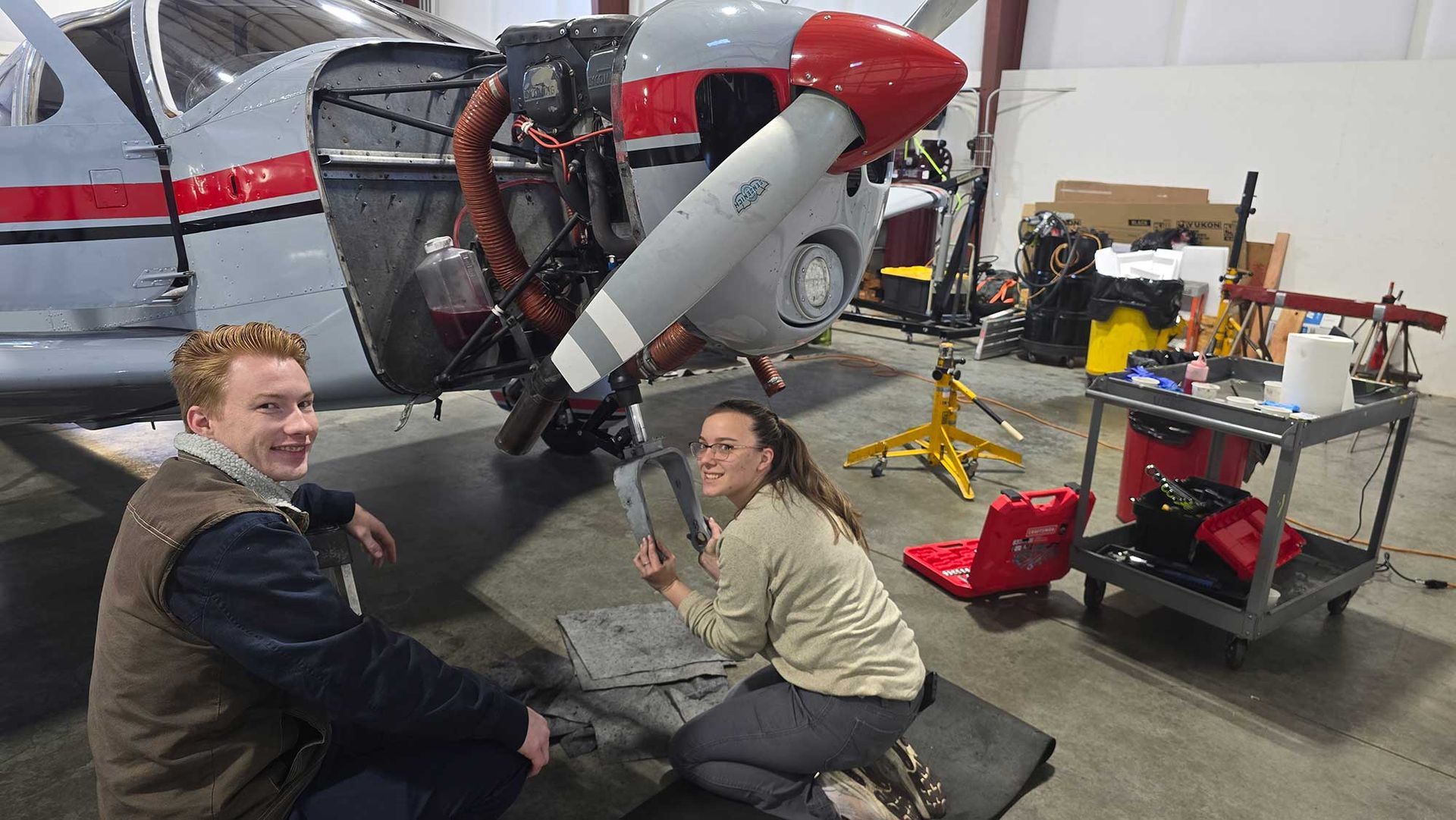 Two people working on a plane engine in a hangar. One smiles at the camera, the other crouches with a part.