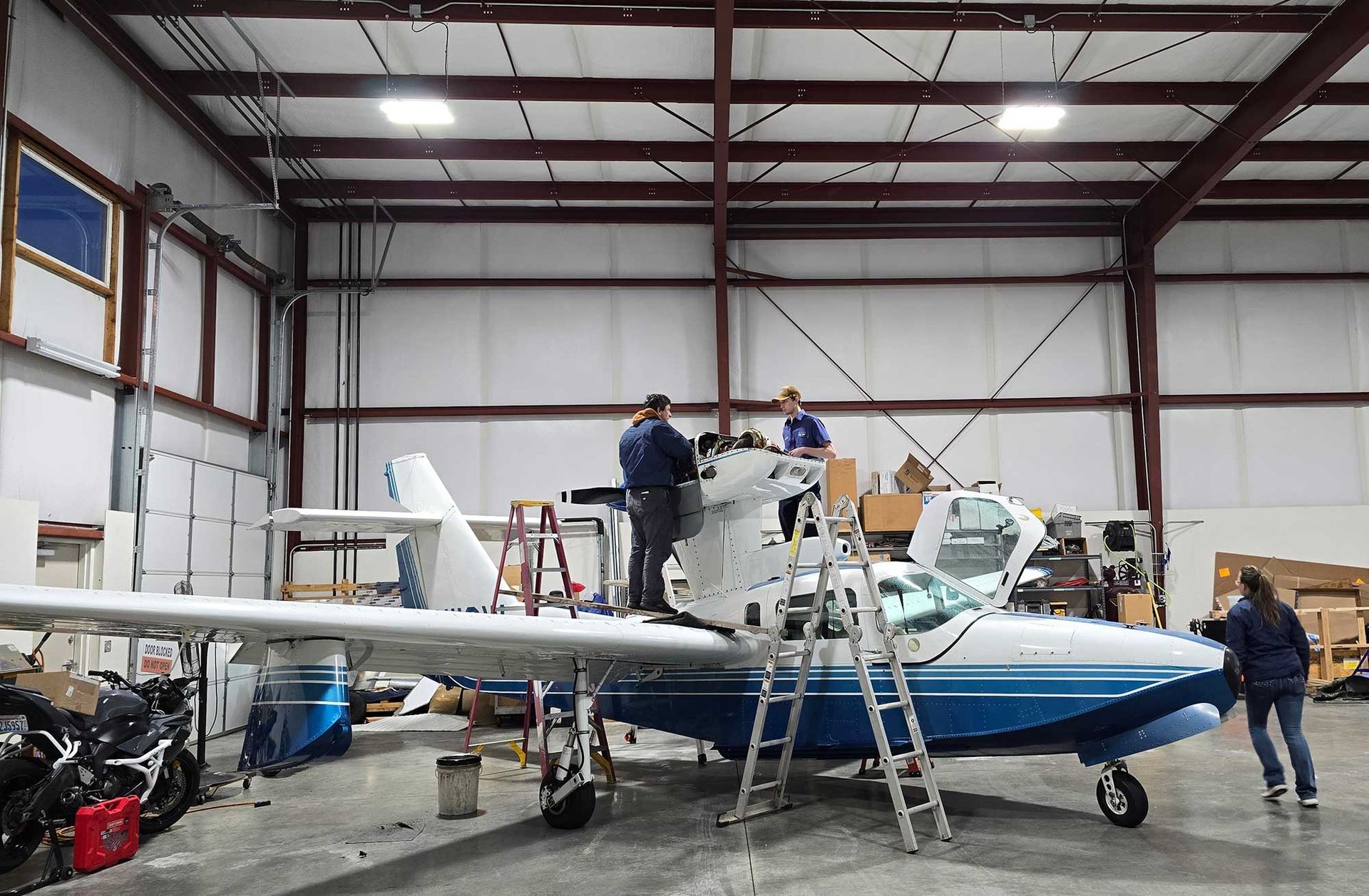 People working on a seaplane inside a hangar. The plane is white and blue, with ladders and tools around it.