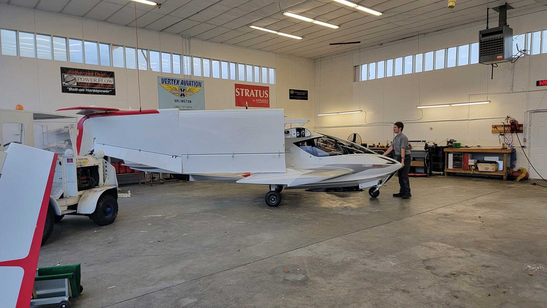 An airplane inside a hangar, wings extended. A person stands nearby, examining it. White exterior, red accents.