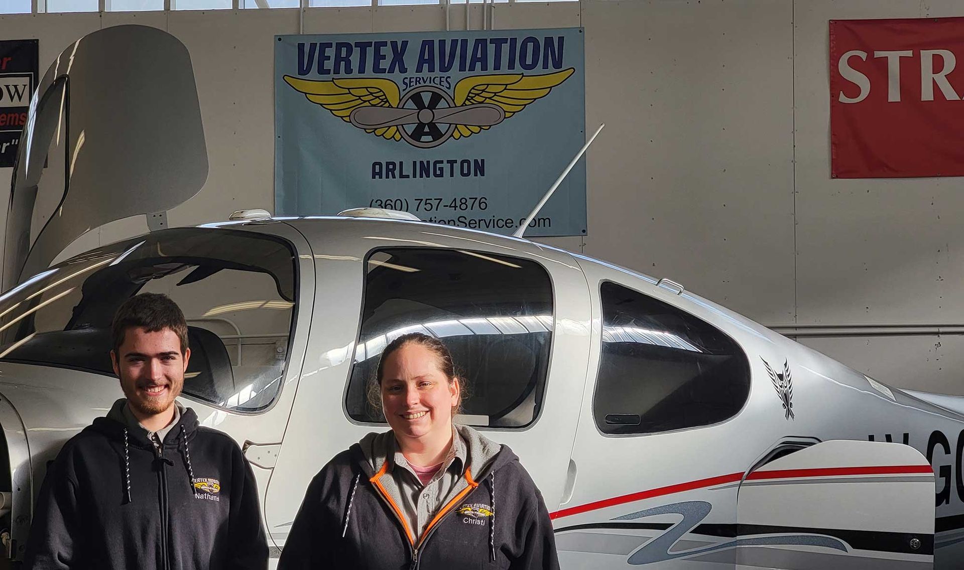 Two people stand in front of a silver airplane inside a hangar. 