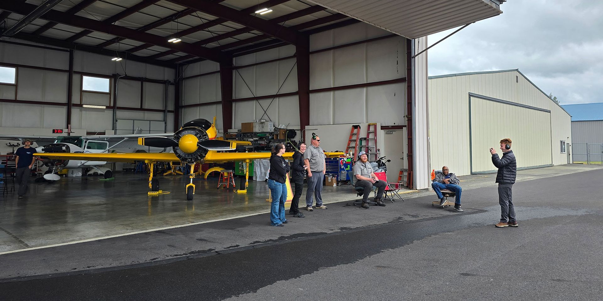 People stand in front of a yellow airplane inside an open hangar. A person takes a photo.