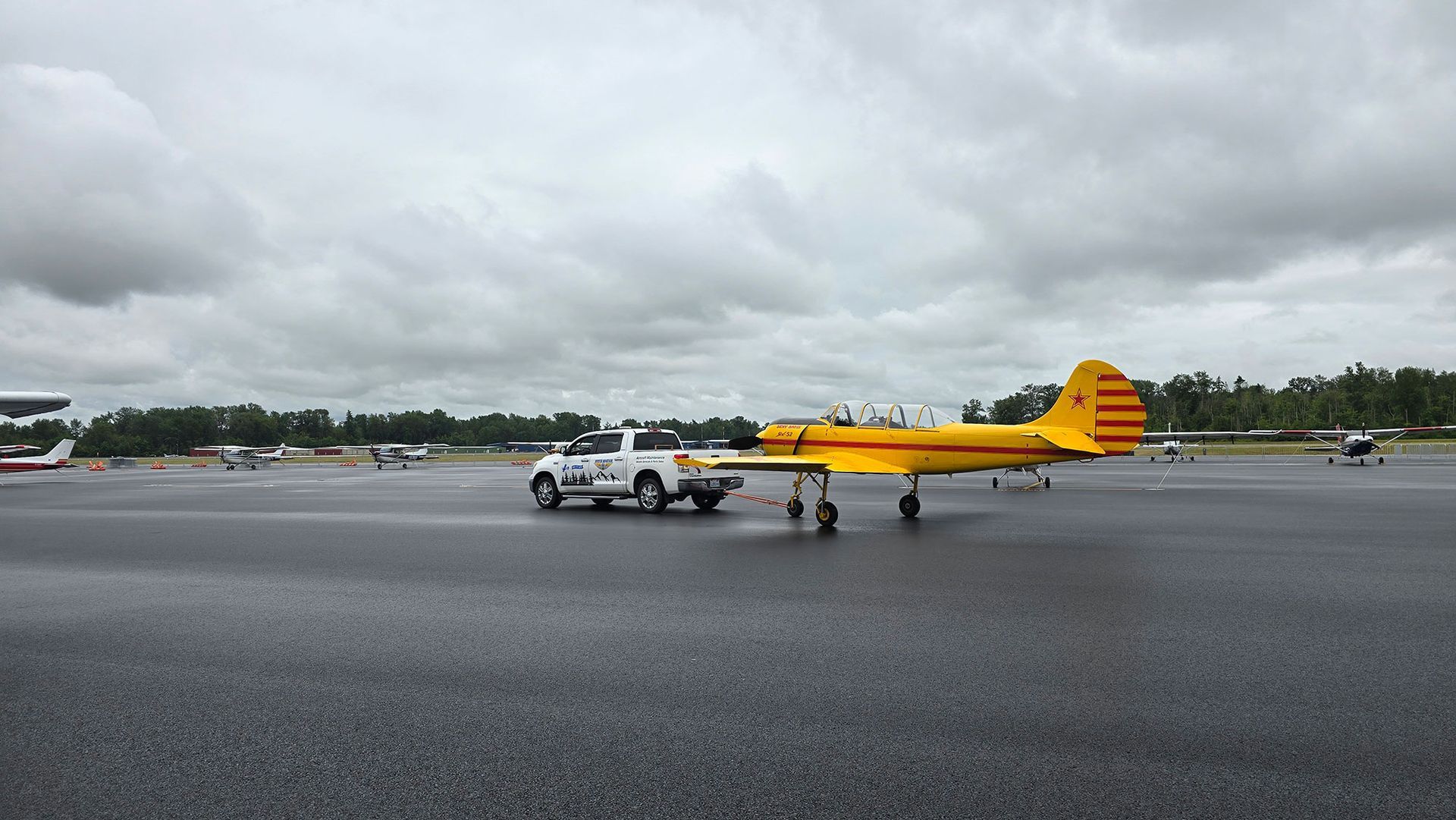 Yellow plane on a wet tarmac, with a white truck. Overcast sky.