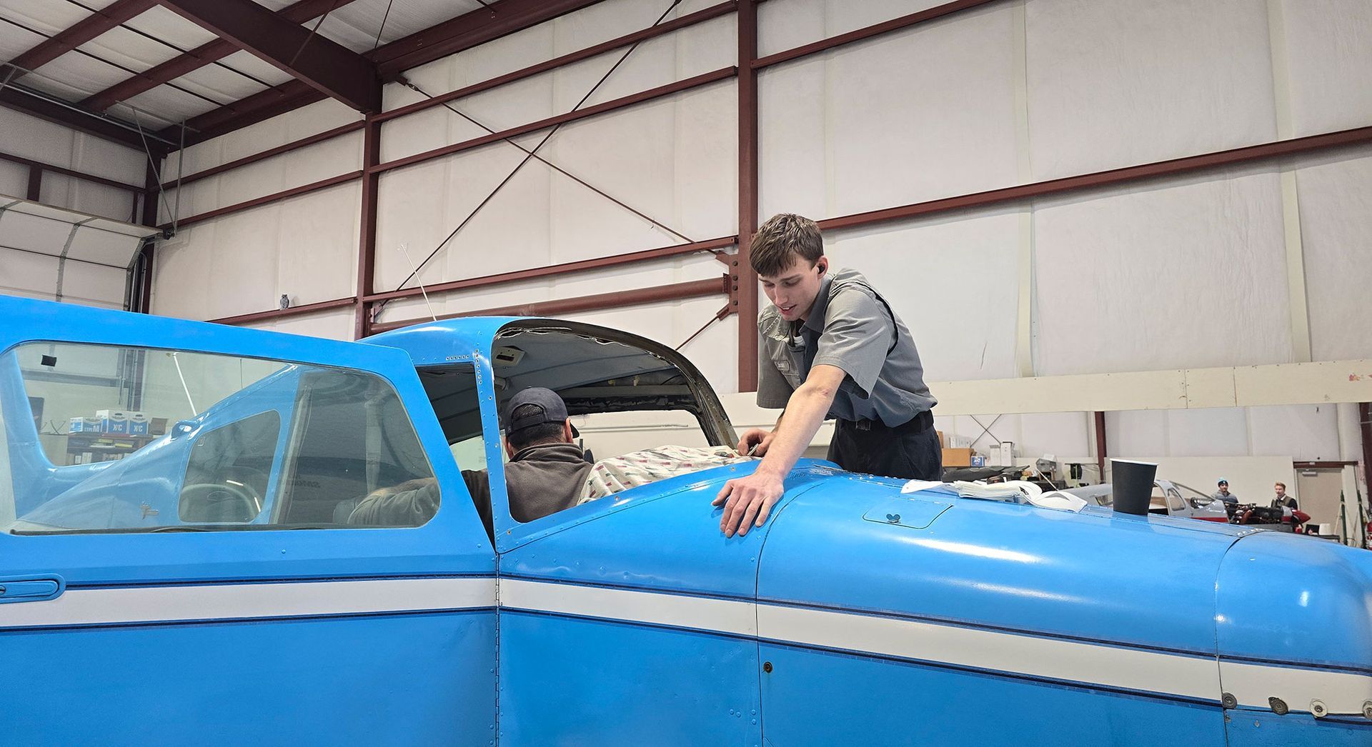 Two people working on a blue airplane in a hangar. One leans over the hood.