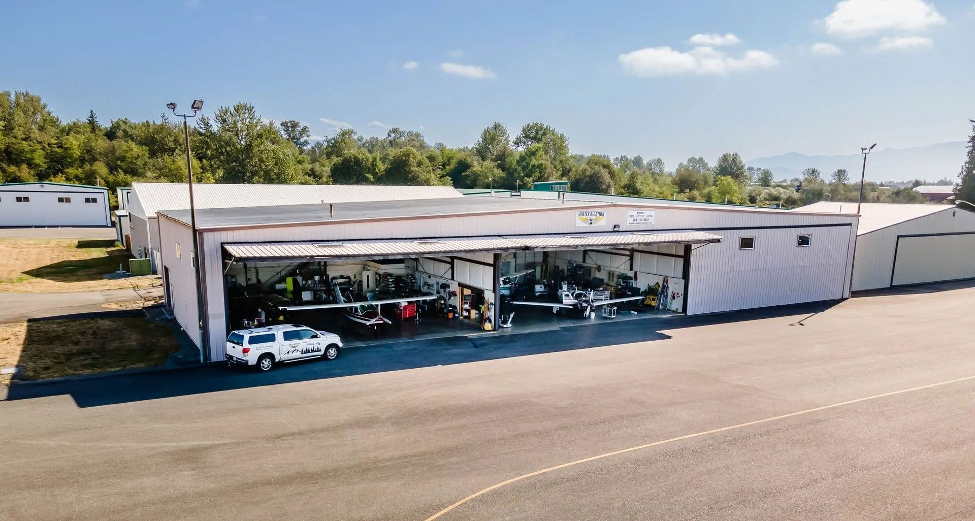 Airport hangar with open doors, showing parked airplanes, vehicles, and equipment; sunny day.