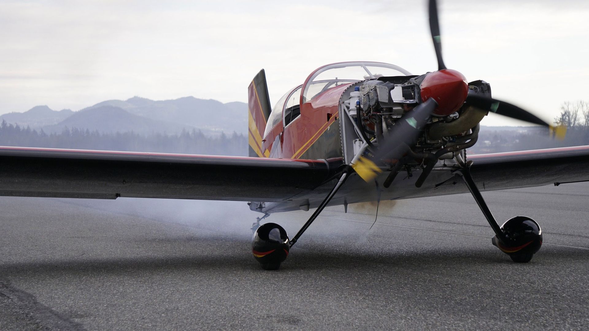 Red and yellow airplane with spinning propeller on a runway, mountains in the background.