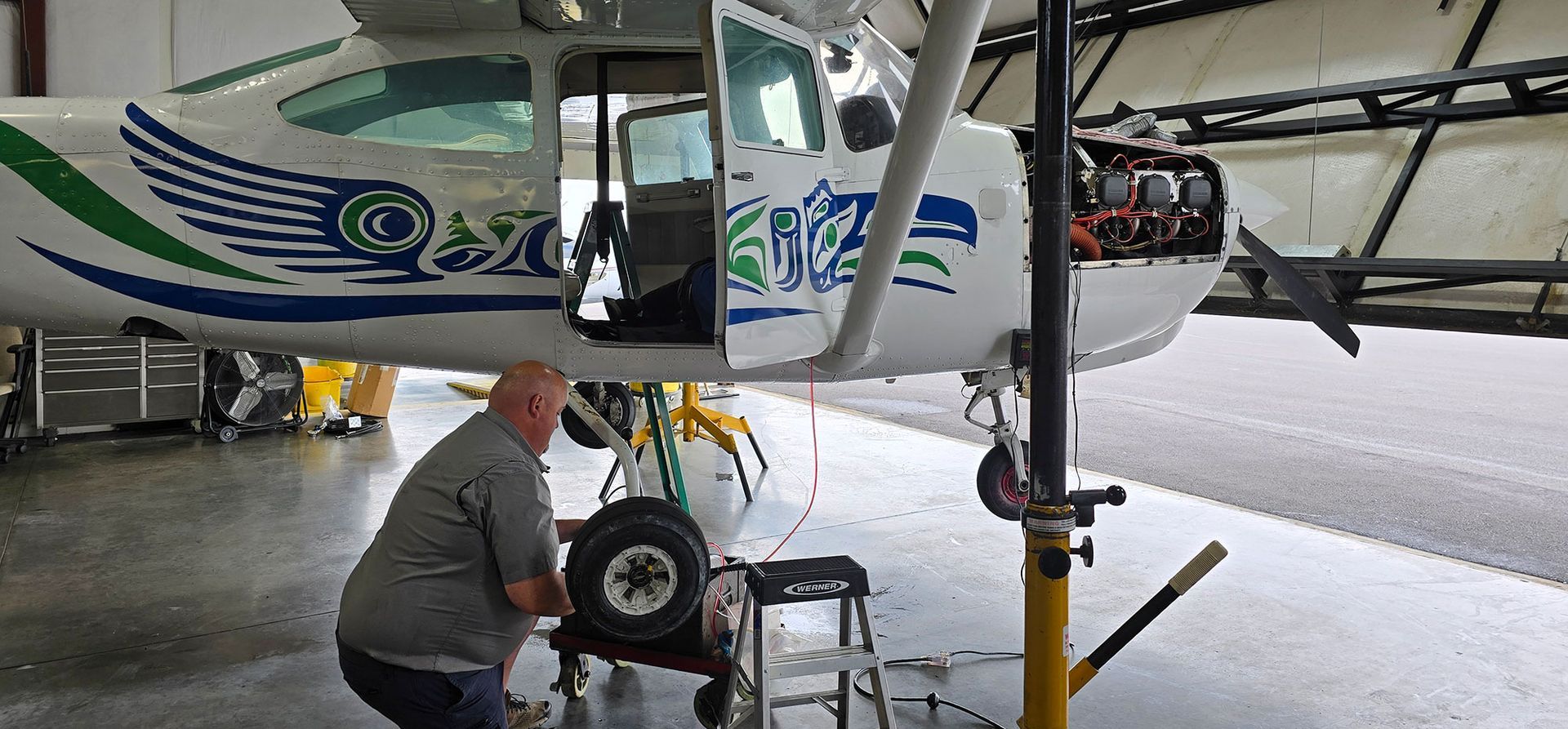 A man is working on an airplane's wheel in a hangar. The plane has blue and green graphics.