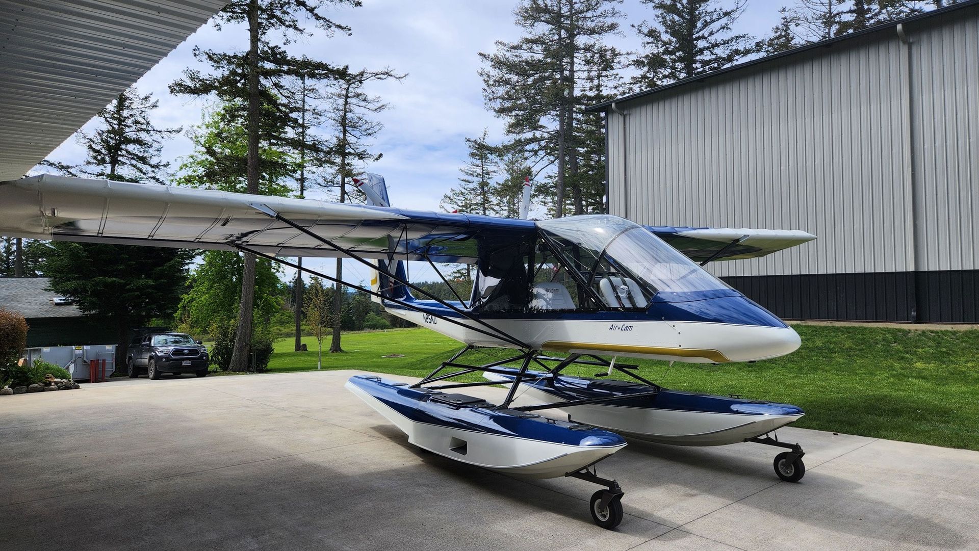 Seaplane parked on a concrete surface, with blue and white pontoons and fuselage. Green trees and a building in background.