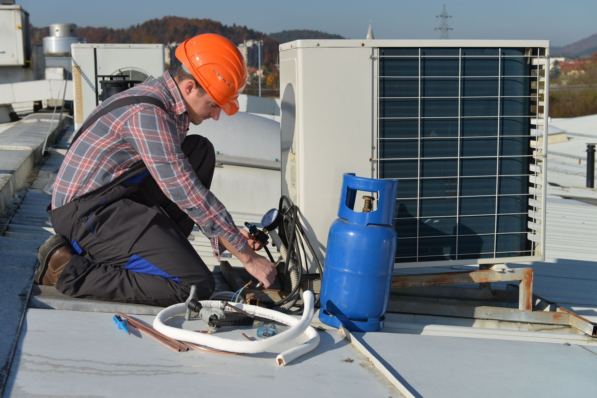 HVAC technician, on rooftop, examines air conditioning unit with gauges; sunny day.