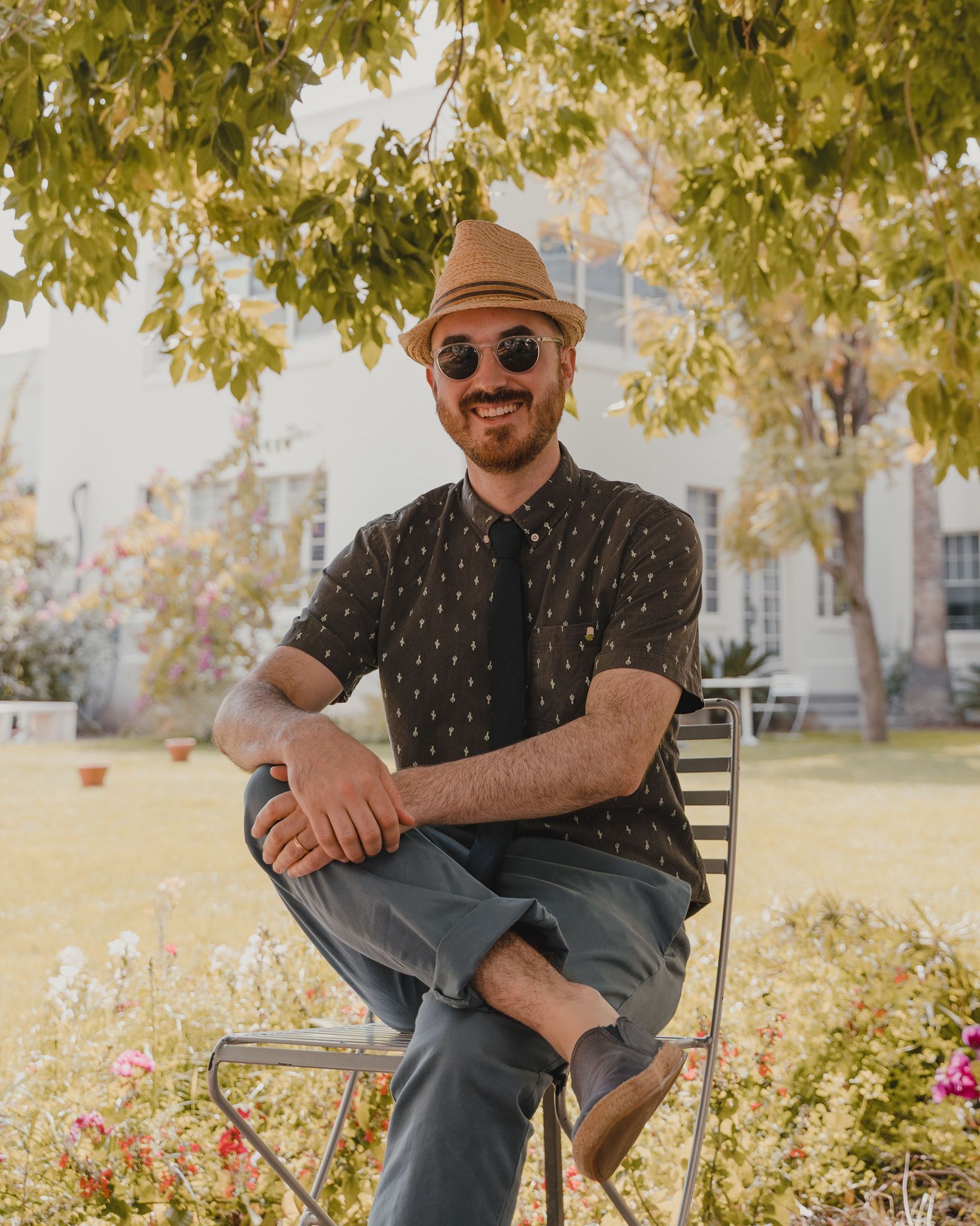 A man wearing a hat and sunglasses is sitting on a chair under a tree.