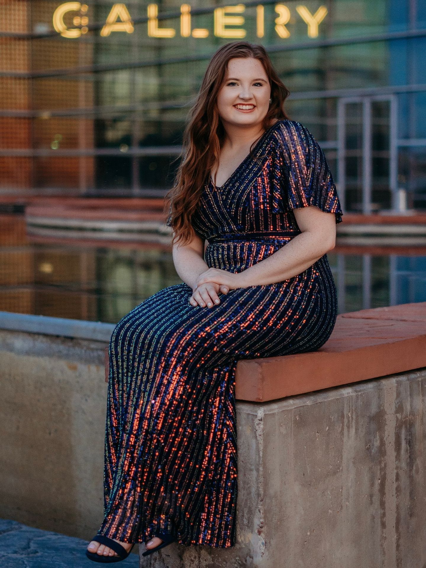 A woman in a long dress is sitting on a wall in front of a building.