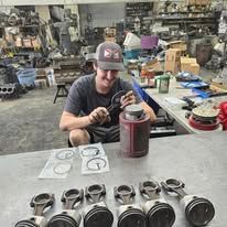 A man is sitting at a table in a garage working on pistons.