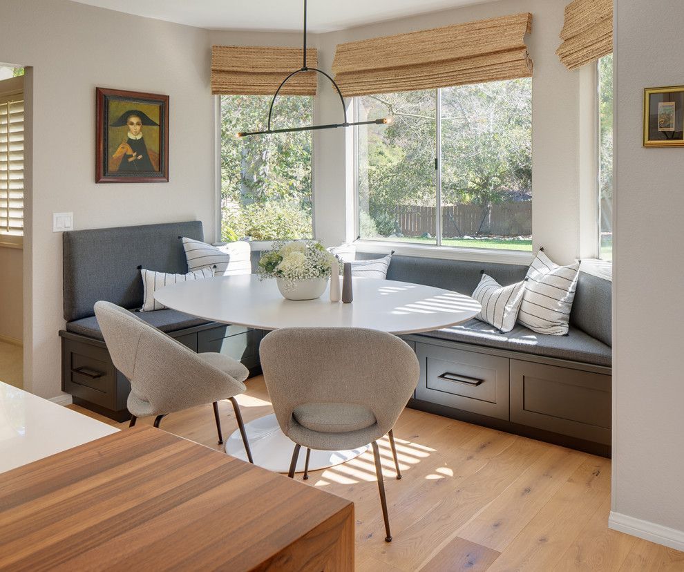 Bright breakfast nook with round table, gray benches, and large windows with woven shades.