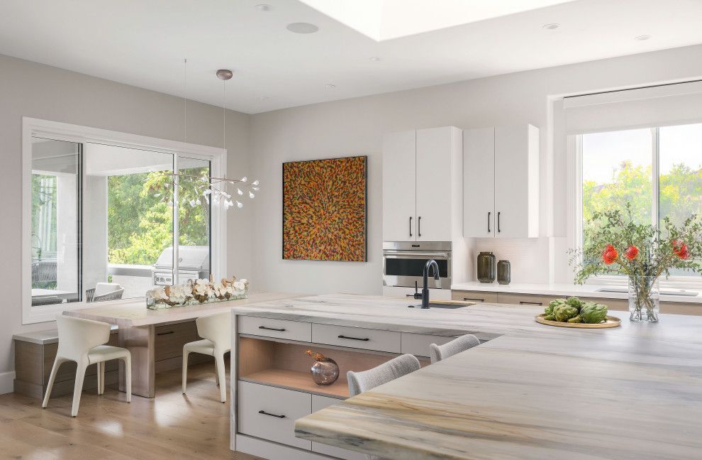 Bright modern white kitchen with island, dining nook, and large windows overlooking greenery