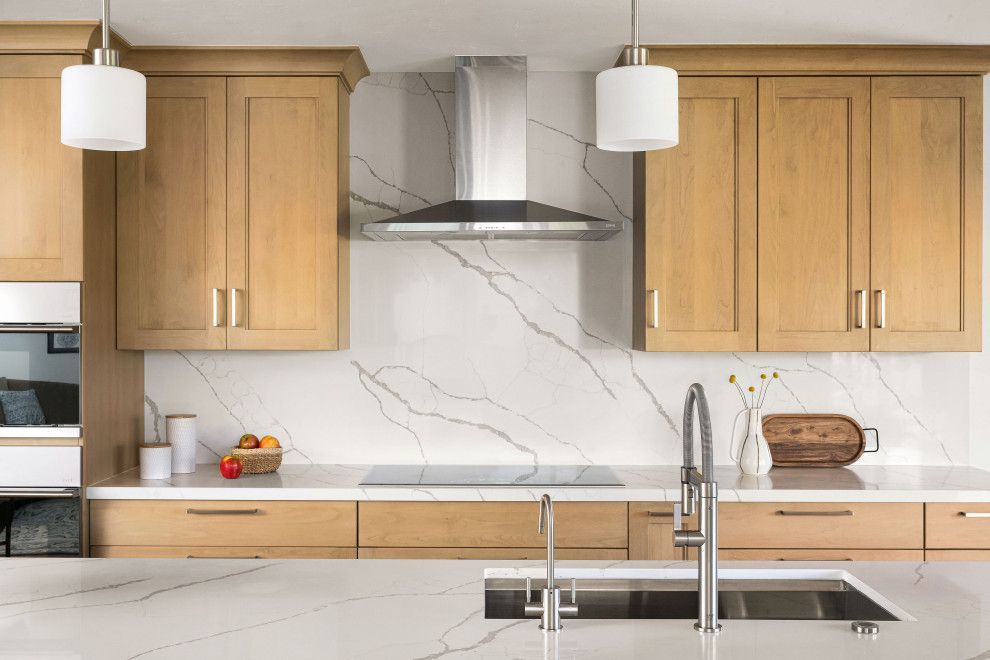 Modern kitchen with light wood cabinets, marble backsplash, stainless range hood, sink, and pendant lights