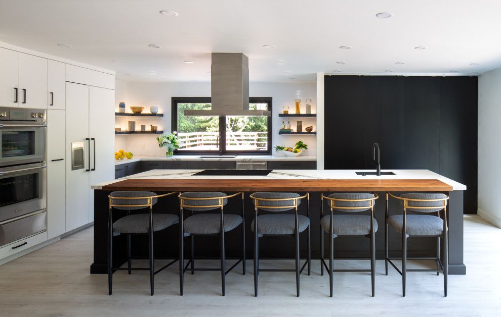 Modern kitchen with large island, black stools, white cabinets, and window shelves with plants