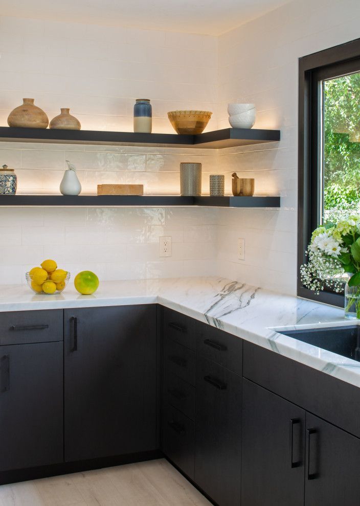 Modern kitchen with dark cabinets, white countertops, open shelves, and a window overlooking greenery