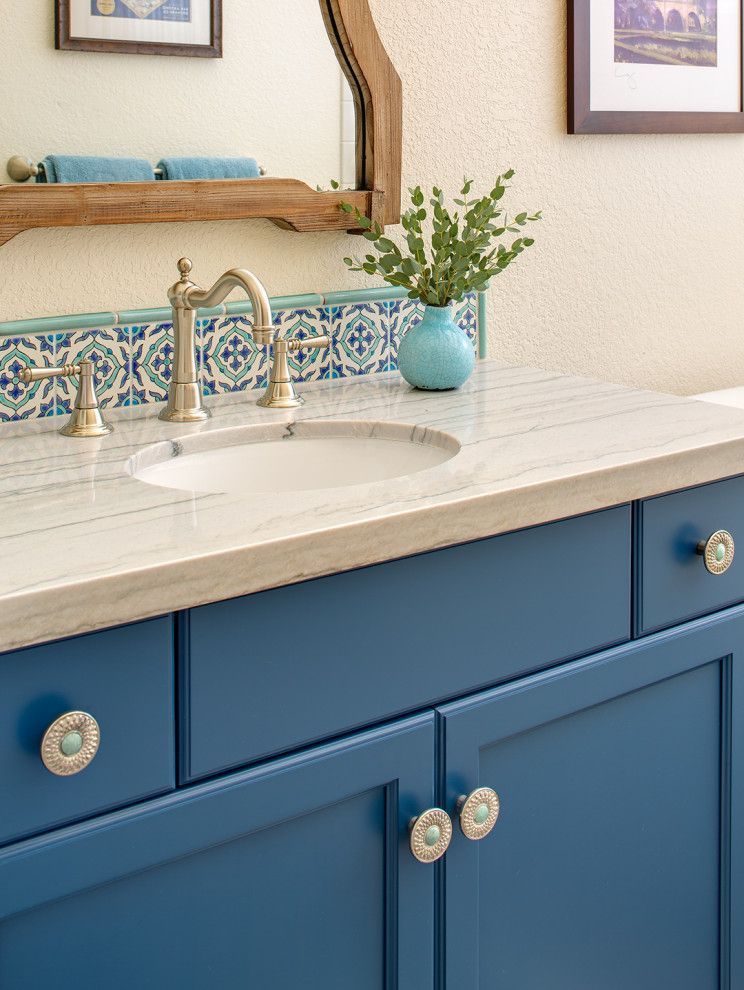 Blue bathroom vanity with white countertop, round sink, brass faucet, and small vase of greenery