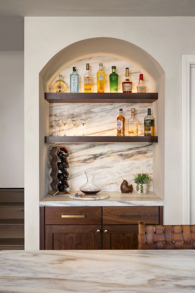 Built-in arched kitchen nook with wooden shelves, liquor bottles, and marble countertop and backsplash