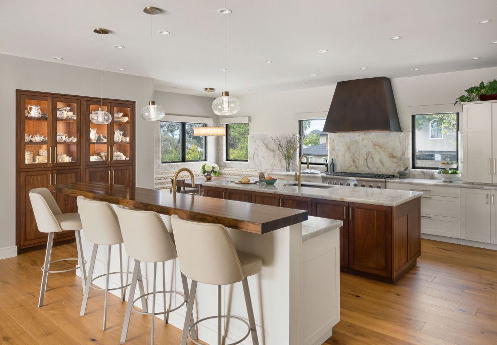 Modern kitchen with white island stools, wooden cabinets, large windows, and a stone range hood