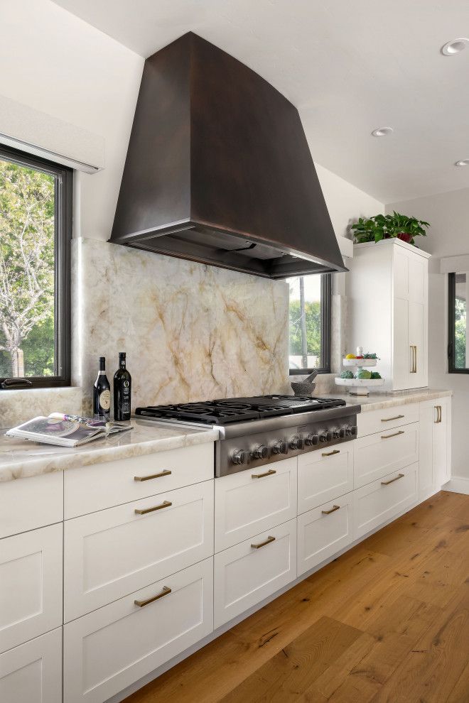 Modern white kitchen with stainless stove, dark range hood, marble backsplash, and wood floor