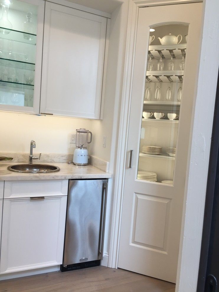 Small white kitchenette with sink, mini fridge, mirror, and glass-front cabinet of dishes.