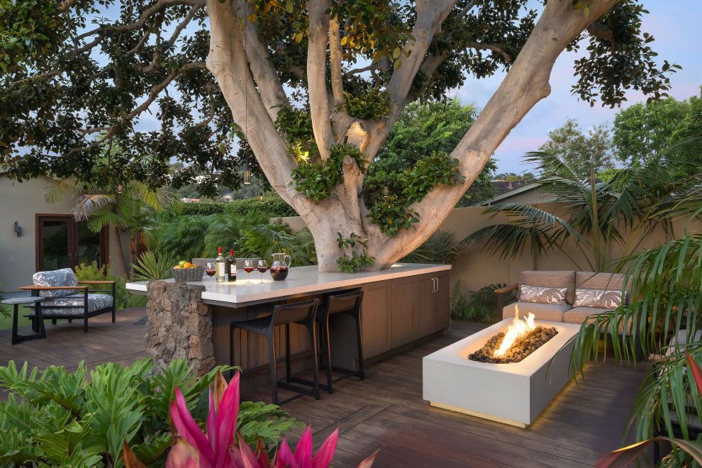 Outdoor patio with a large tree, bar counter, seating, and a fire pit surrounded by greenery.