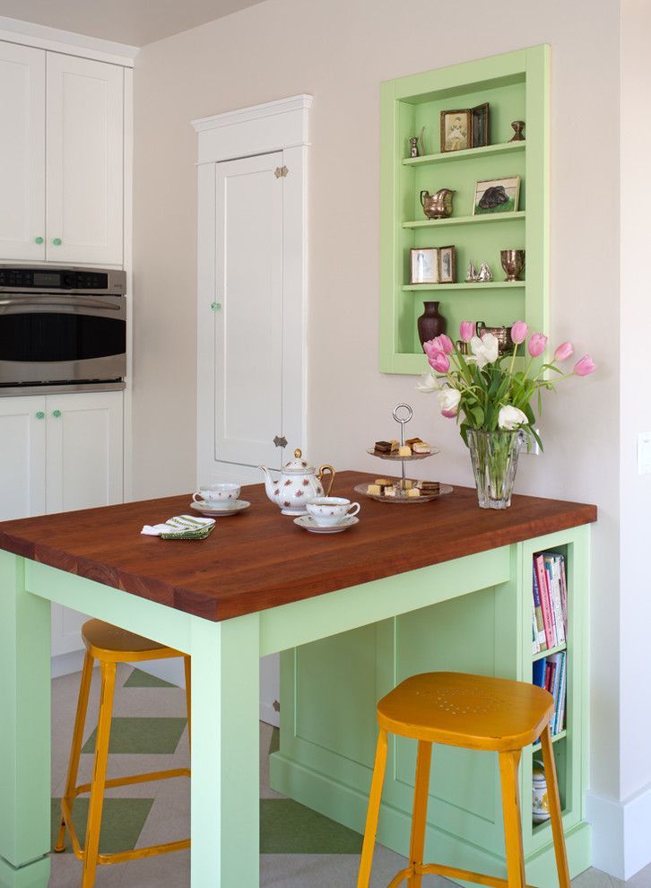 Mint-green kitchen island with wooden countertop, yellow stools, tea set, and flowers in a bright corner.