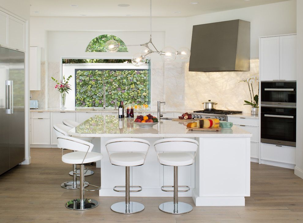 Bright white modern kitchen with island, bar stools, and stainless appliances, lit by a large window.