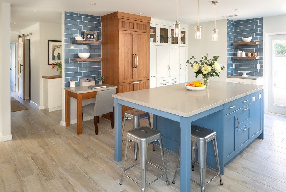 Bright kitchen with blue island, wooden cabinets, stools, and a small dining table by a blue-tiled wall.