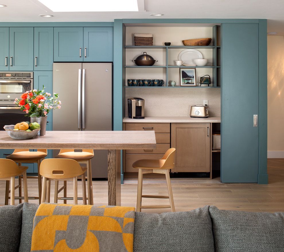 Modern kitchen with teal cabinets, stainless fridge, island stools, and built-in shelves near a gray sofa.